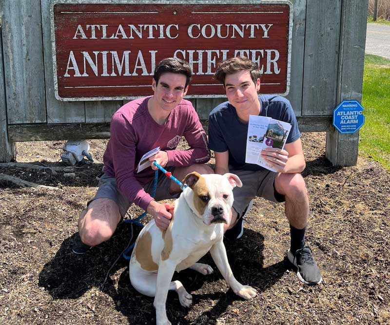 Two young men holding the pamphlets in front of the Animal Shelter Sign with a dog on a leash.
