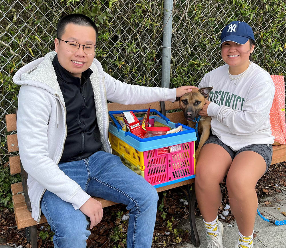 Two Stockton students, one male, one female, site on a bench petting a dog that is sitting on the bench next to a crate of donated items.