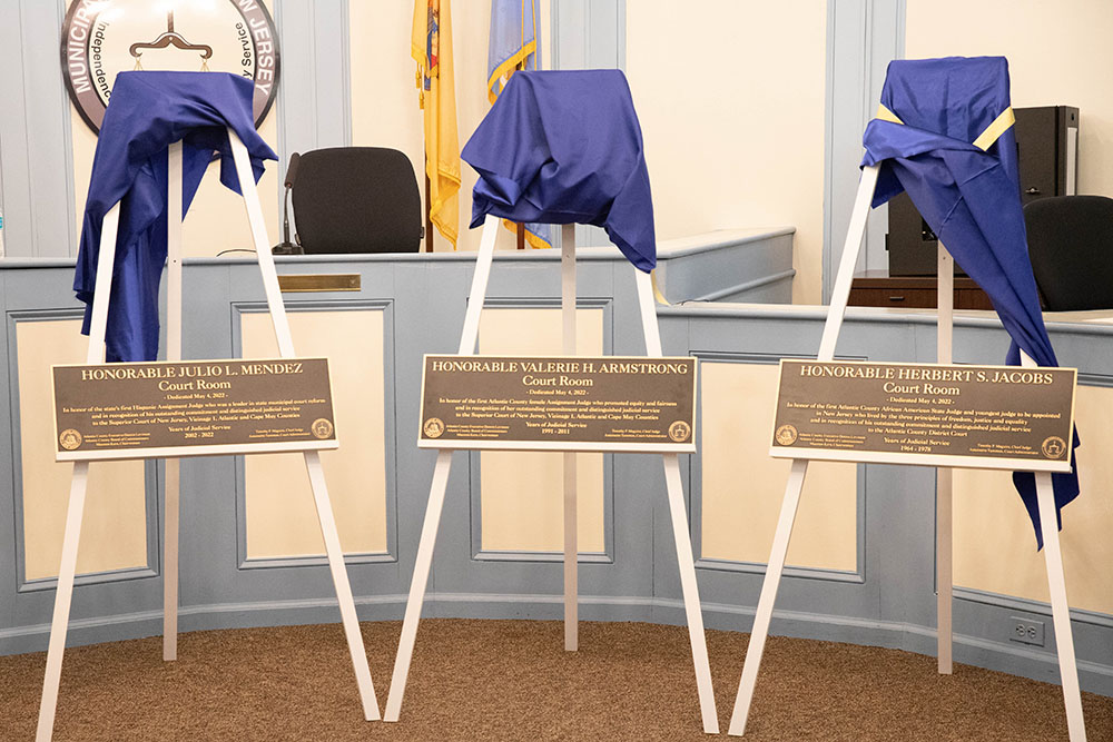 Image shows 3 white easels holding the bronze plaques that will be displayed in the court rooms naming the rooms for the honorees.  Plaques are bronze in color and with white lettering.